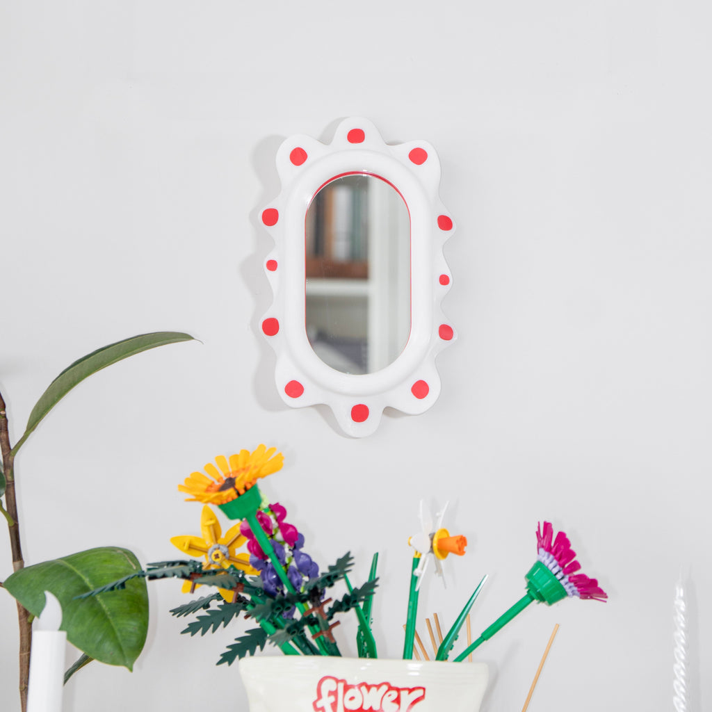 Decorative mirror with red polka dots on a white wall, next to a colorful flower pot with flowers.