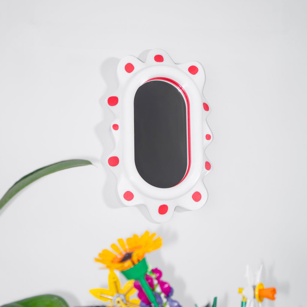 Decorative mirror with red polka dots on a white wall, next to a colorful flower pot with flowers.