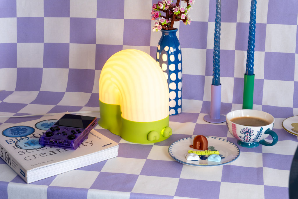 A glowing snail-shaped lamp with a white dome and bright lime-green base on a lavender checkered tablecloth, styled with blue twisted candles, a spotted blue vase of pink Lego flowers