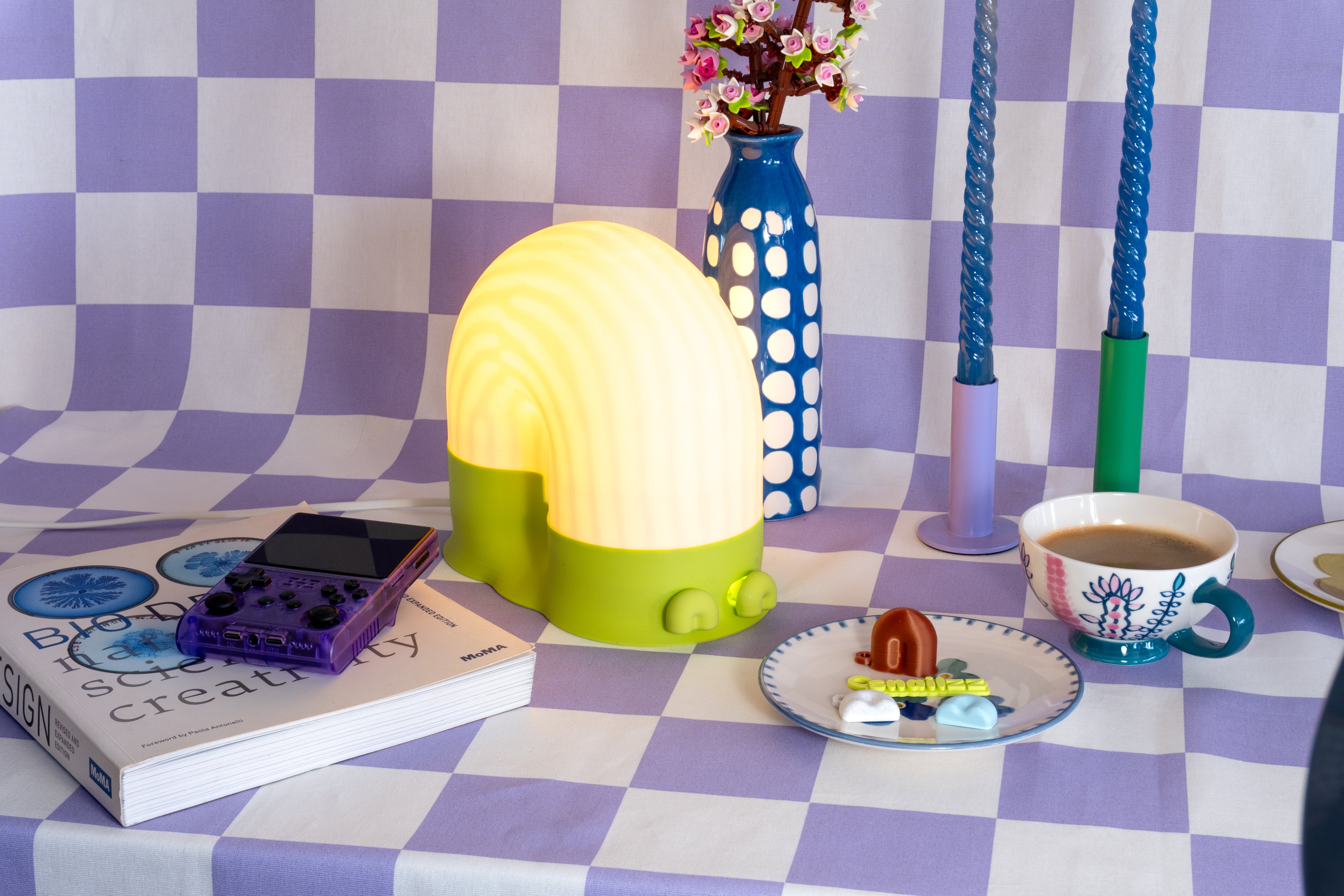 A glowing snail-shaped lamp with a white dome and bright lime-green base on a lavender checkered tablecloth, styled with blue twisted candles, a spotted blue vase of pink Lego flowers