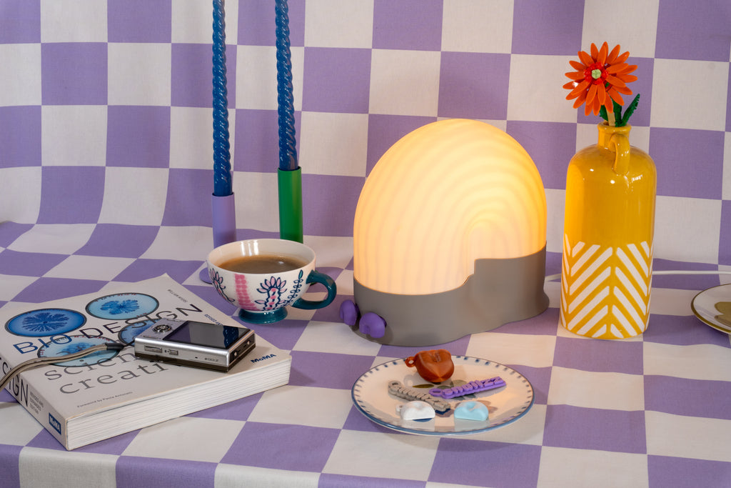A glowing snail-shaped lamp with a grey base and pastel shell placed on a lavender checkered IKEA tablecloth, surrounded by a yellow vase with an orange flower, twisted blue candles on a IKEA candle holder , a patterned teacup, and a MOMA design book.
