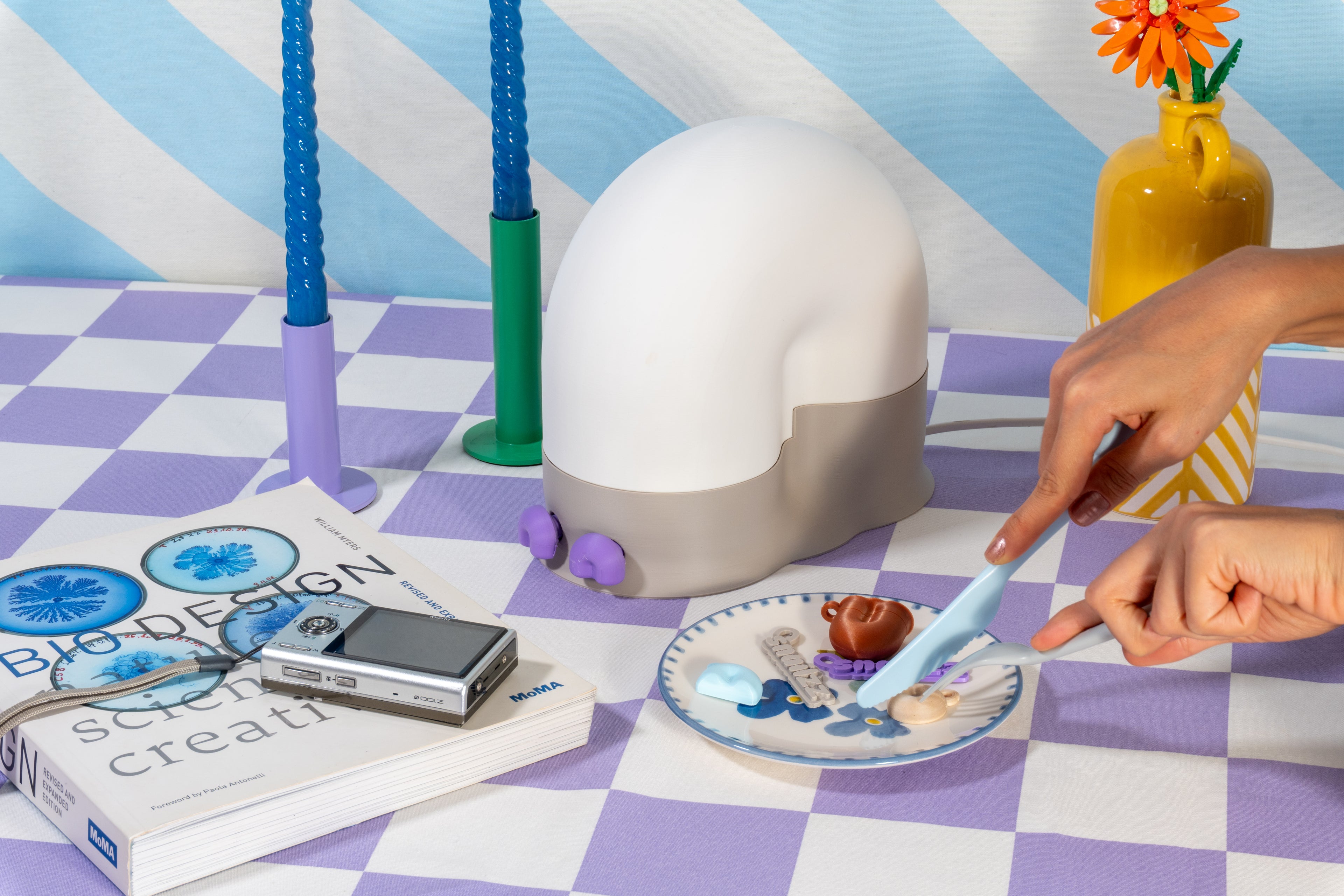 A snail-inspired lamp with a white shell and grey base sits on a lavender checkered IKEA tablecloth as a person playfully uses pastel blue IKEA cutlery on a plate with colorful trinkets, next to a FUJIFILM retro camera, MoMA design book, and decorative candles.