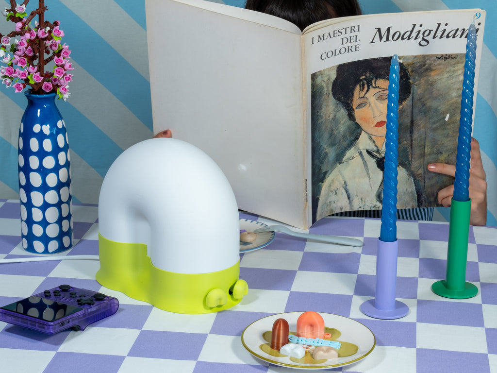 A snail-shaped lamp with a white dome and bright lime-green base on a lavender checkered tablecloth, styled with blue twisted candles, a spotted blue vase of pink flowers, and a person reading a Modigliani art book behind it.