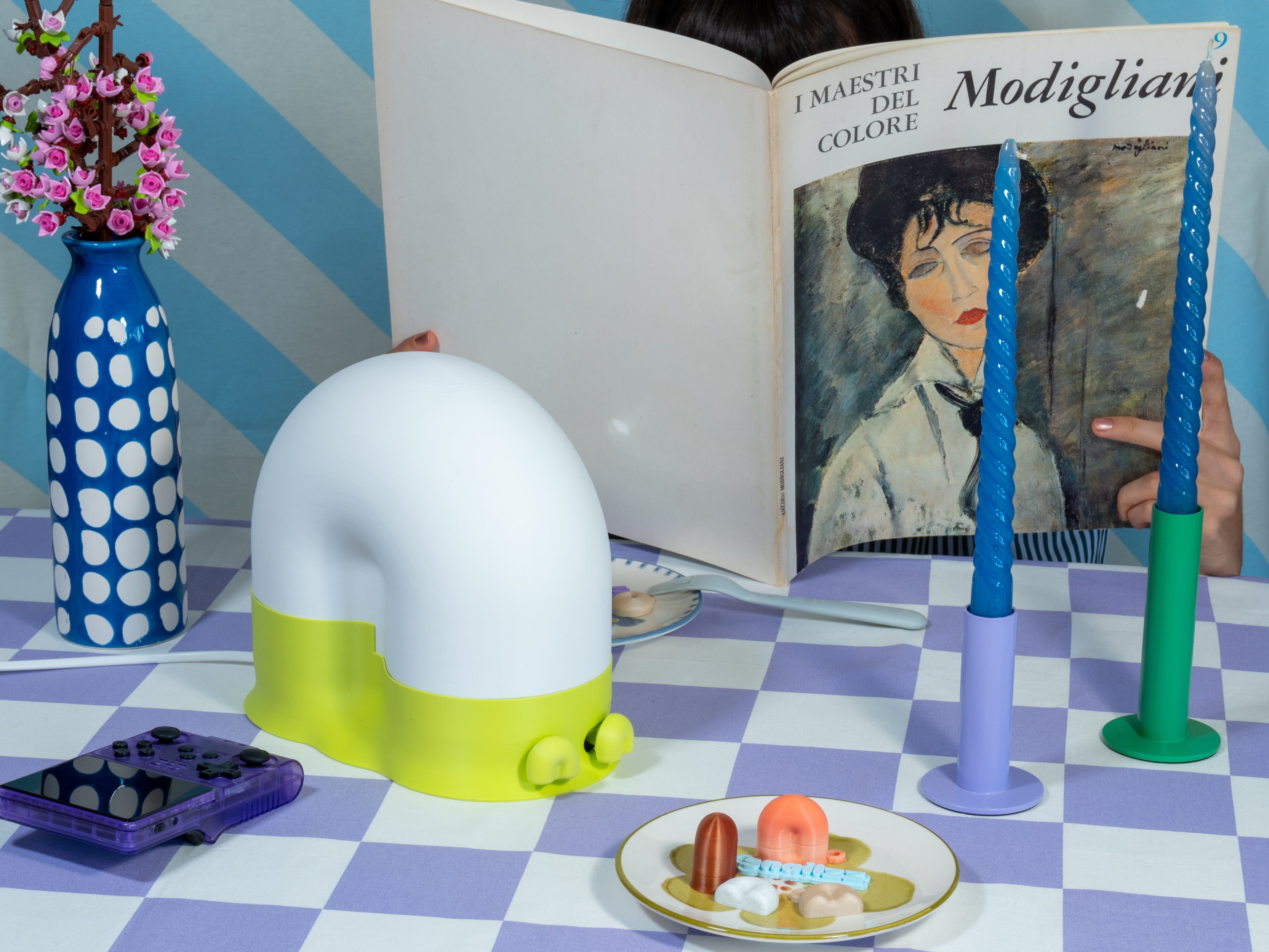 A snail-shaped lamp with a white dome and bright lime-green base on a lavender checkered tablecloth, styled with blue twisted candles, a spotted blue vase of pink flowers, and a person reading a Modigliani art book behind it.