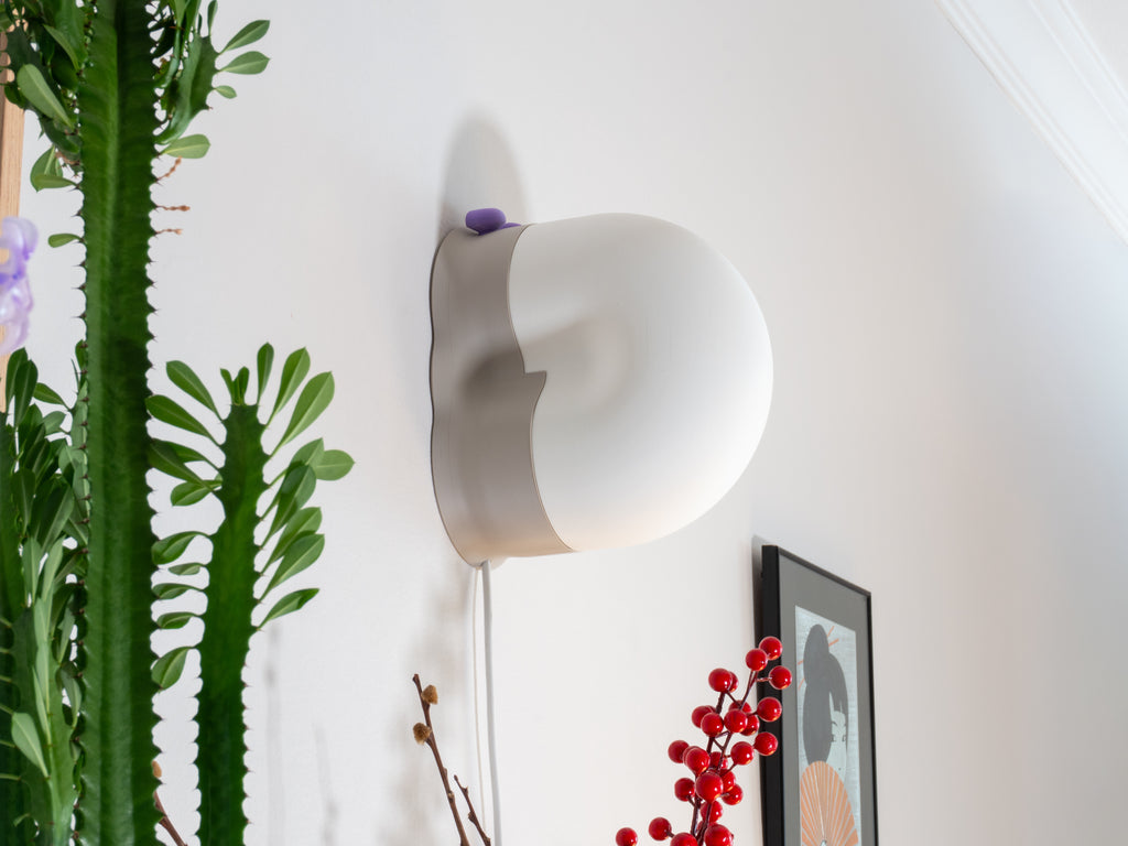 A snail-shaped wall lamp with a soft white shell and grey base mounted on a white wall, styled with green cacti, red berries, and framed artwork in the background.