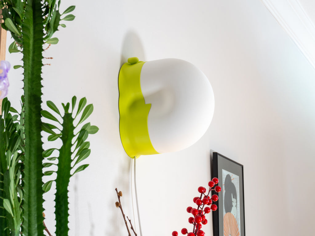 A snail-inspired wall lamp with a white dome shell and vibrant lime-green base mounted on a white wall, surrounded by plants and framed artwork.